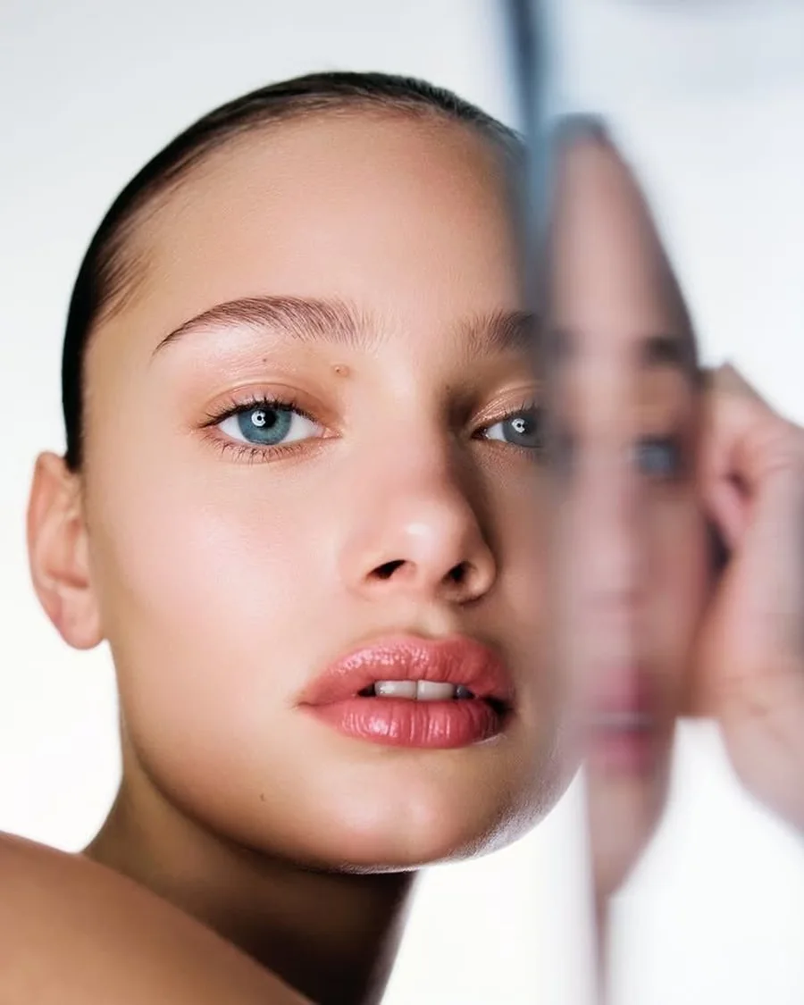 Close-up portrait of a woman with dark hair and glossy lips beside a mirror