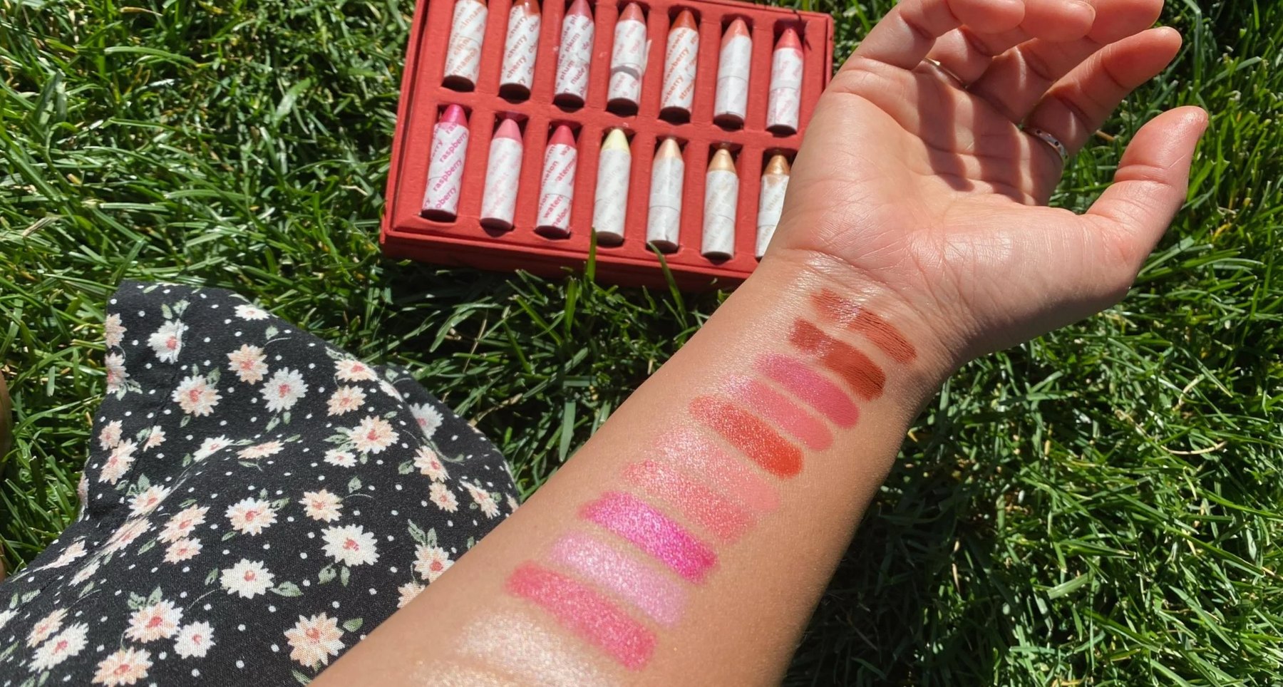 Forearm with makeup swatches beside a tray of Balmie crayons on grass.