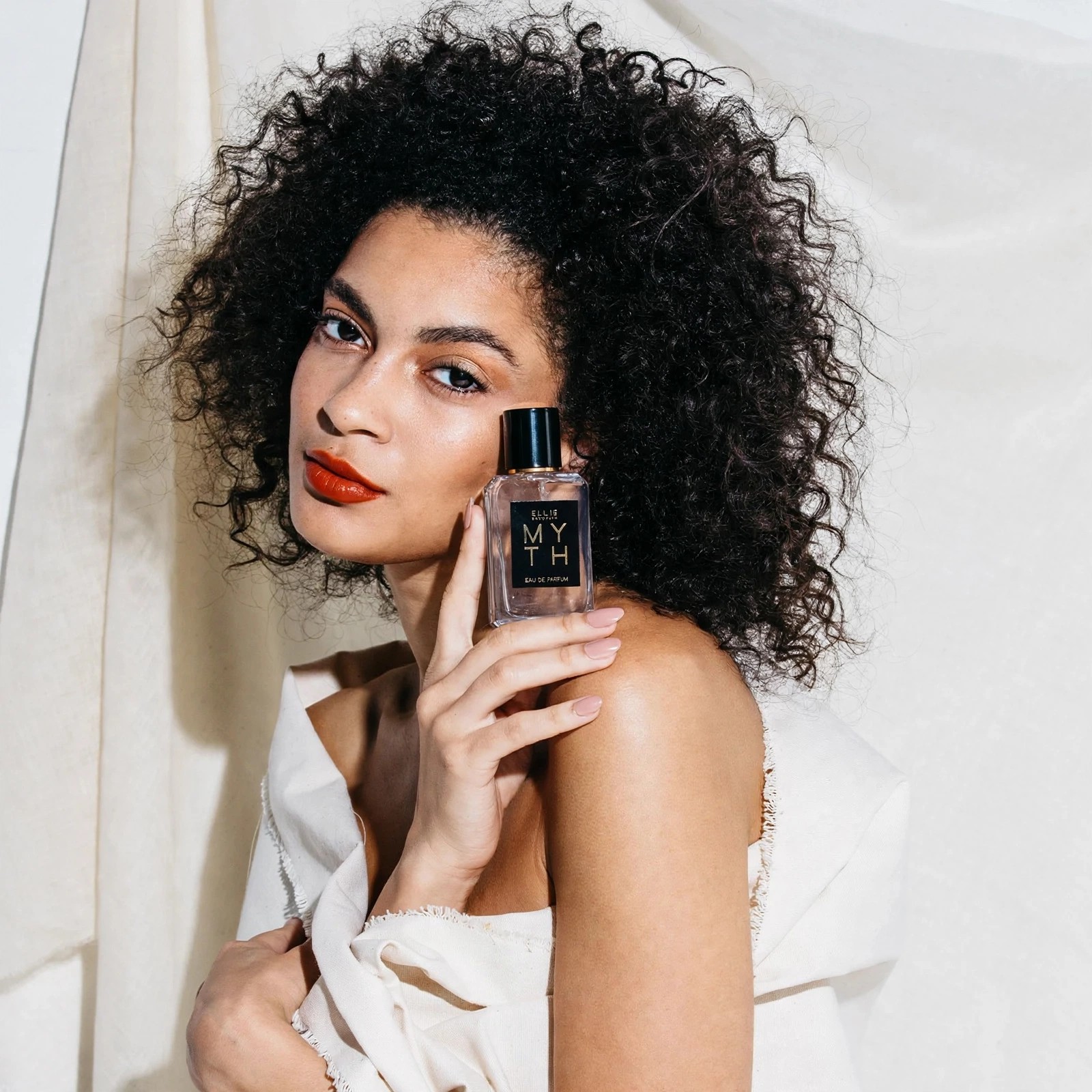 Woman with curly hair holding Myth perfume bottle near shoulder.