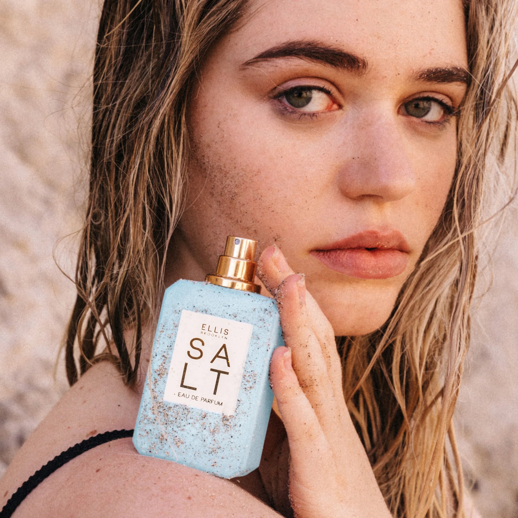Woman holding Salt perfume bottle against sandy cheek outdoors.