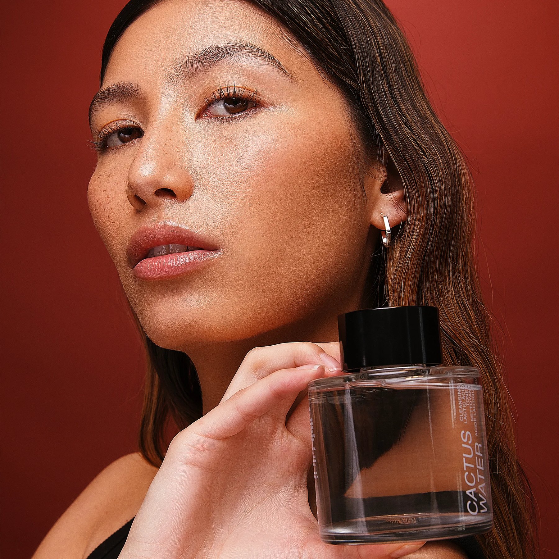 Woman with freckles holding Cactus Water bottle against red background.