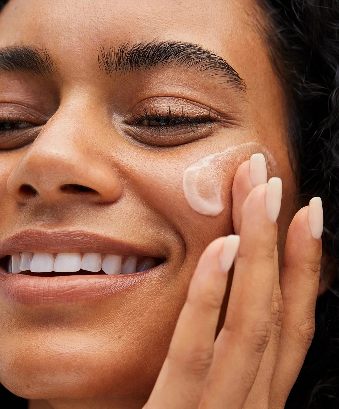Woman smiling while blending cream product onto her cheek.